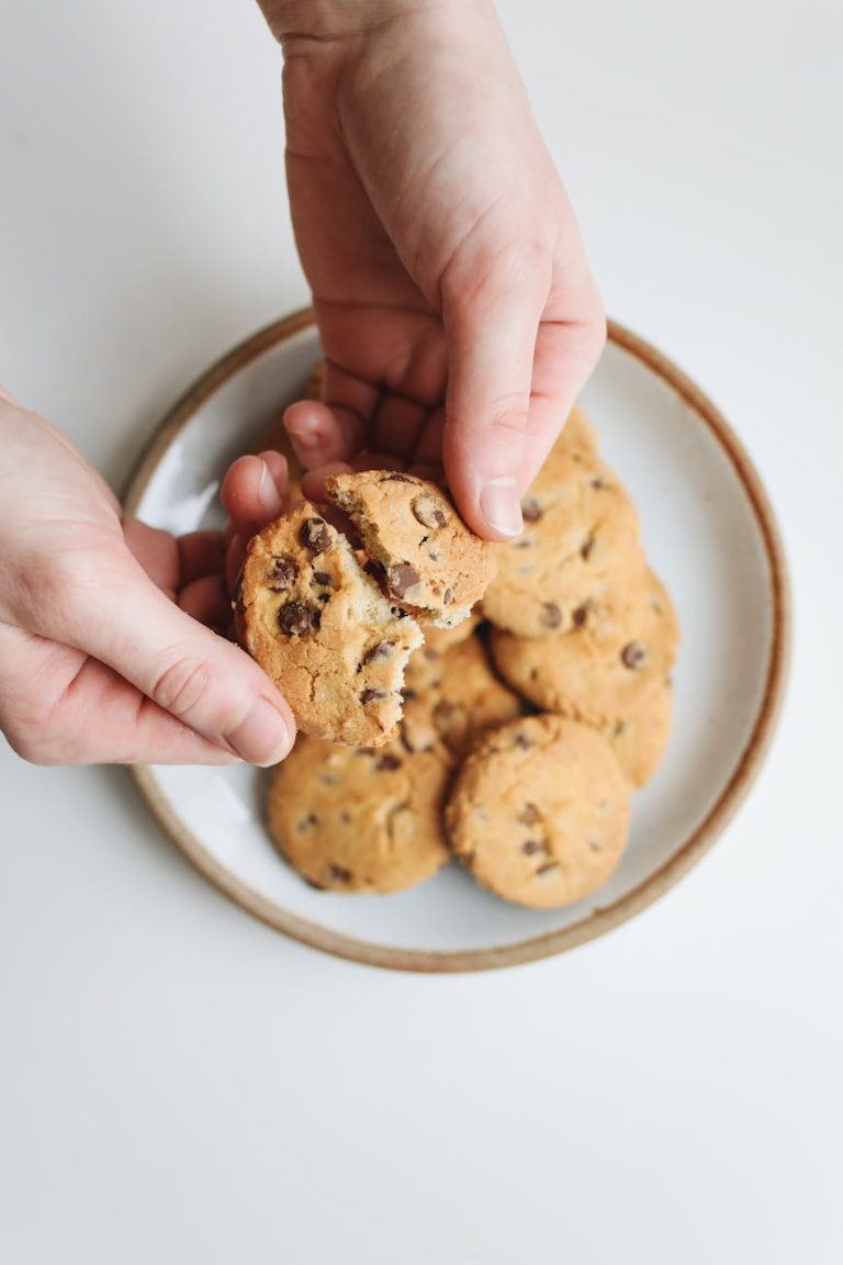Close-up of hands breaking a chocolate chip cookie on a ceramic plate, showcasing homemade dessert.