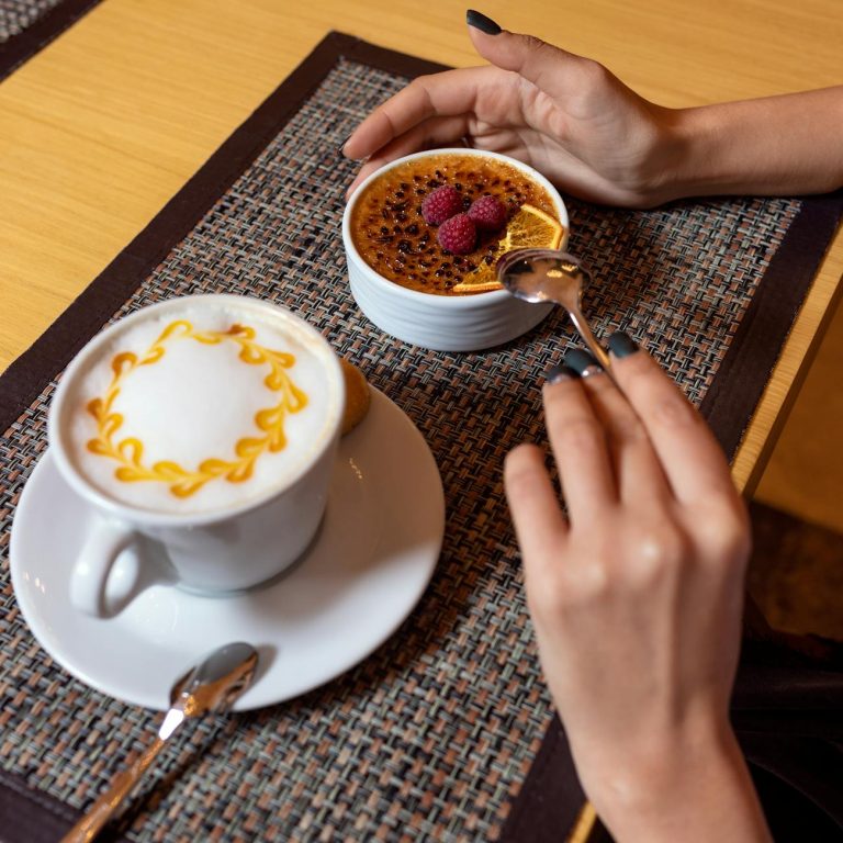 A warm setting with cappuccino and crème brûlée featuring raspberries and a hand holding a spoon.