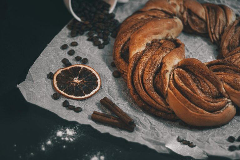 A rustic cinnamon kringle surrounded by coffee beans and dried orange for a cozy bakery setting.