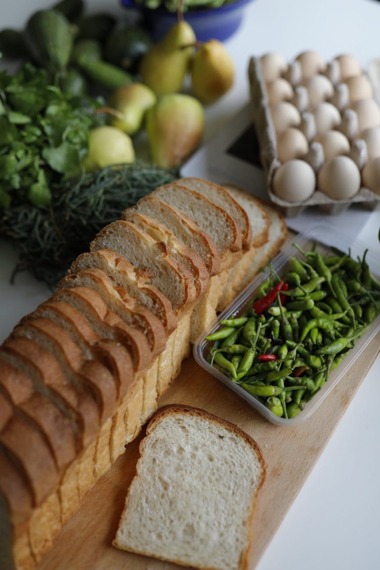 A vertical overhead shot of sliced bread, fresh vegetables, and eggs on a wooden board in a kitchen setting.