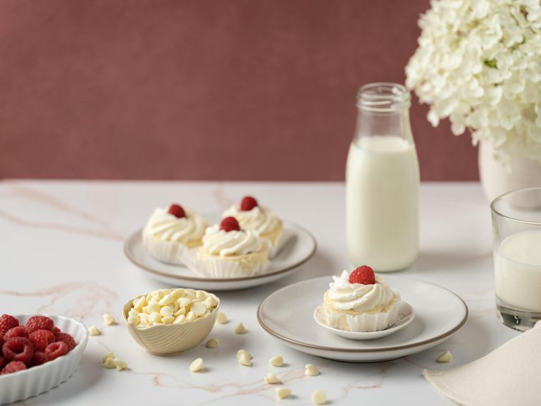 A lovely display of vanilla cupcakes topped with raspberries, accompanied by milk and white chocolate chips.