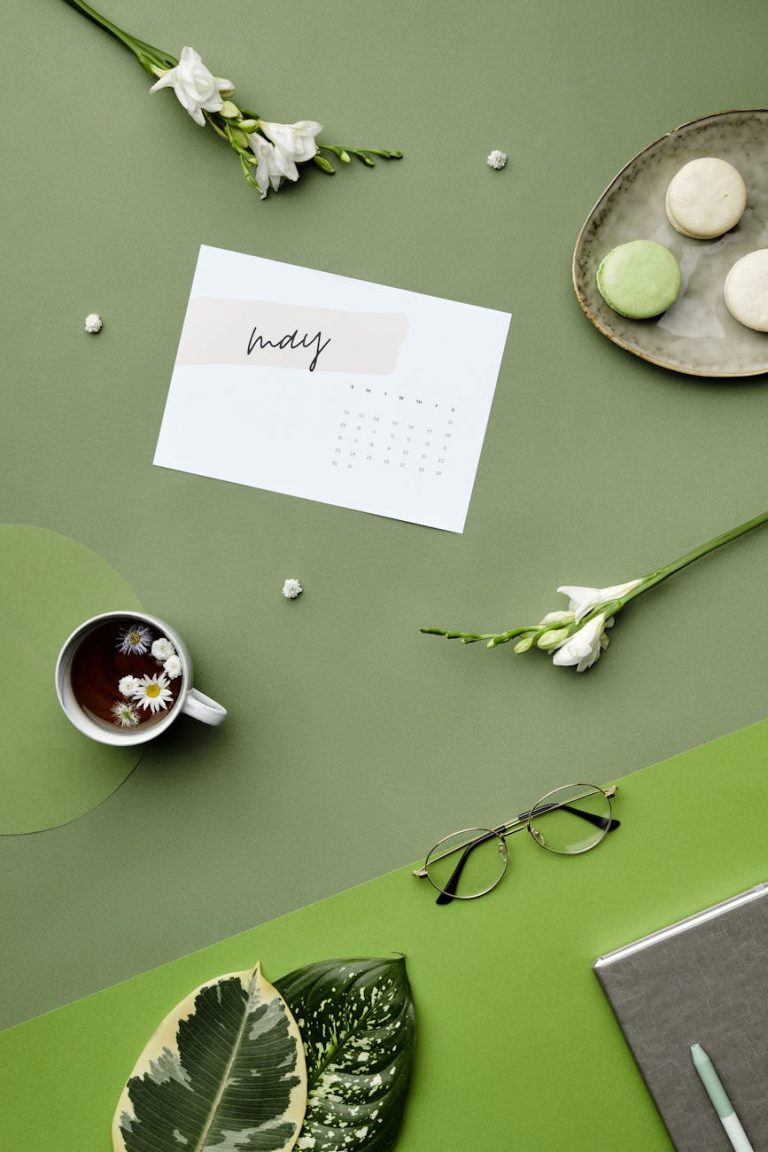 A chic flat lay featuring a May calendar, tea, flowers, and macarons on a green background.